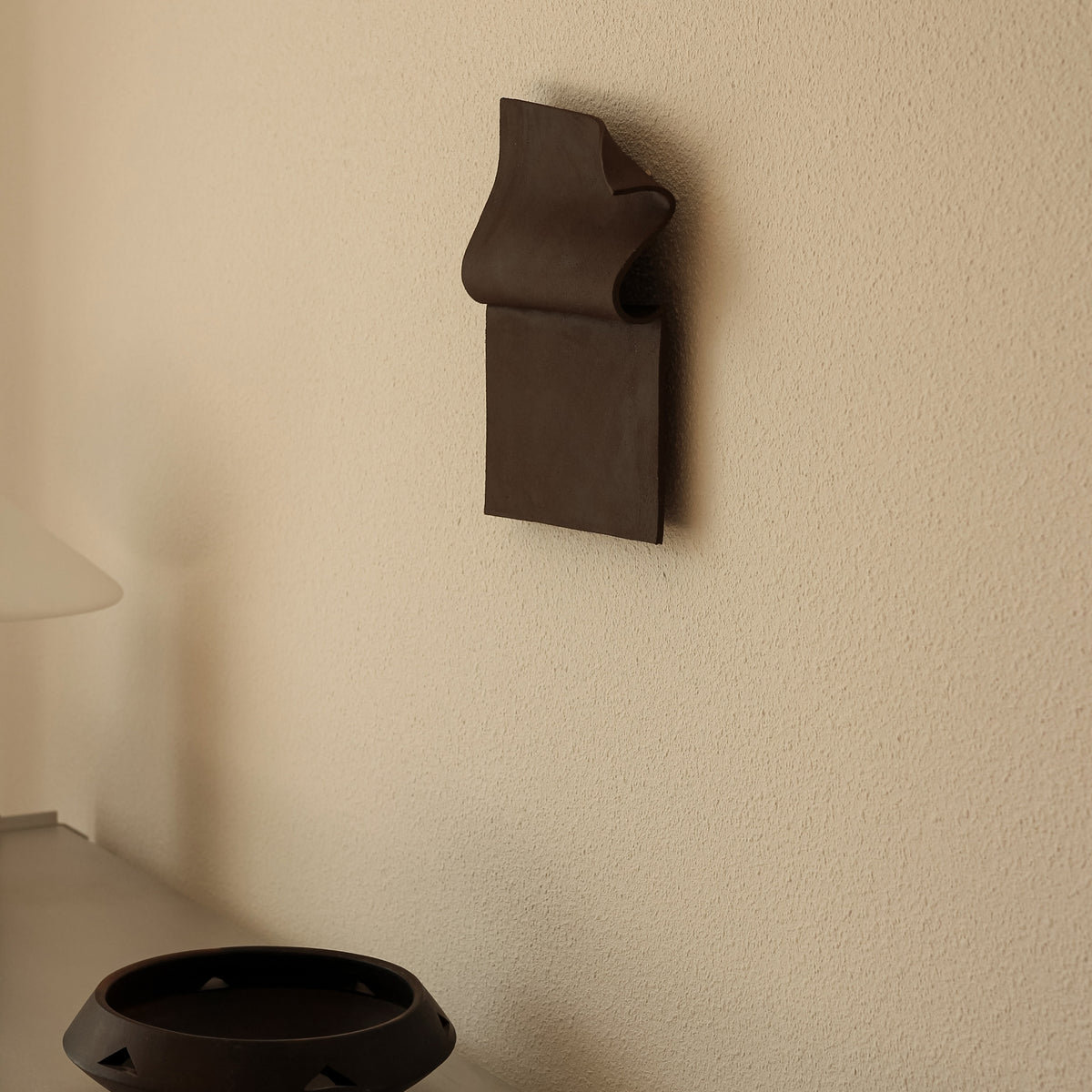 Writings 001 by Studio JOA — Dark Brown folded stoneware wall piece mounted on a plain wall above a ceramic bowl