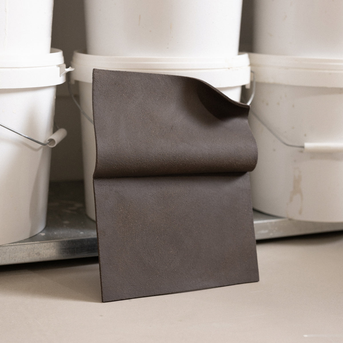 Studio JOA — Dark Brown folded stoneware wall piece standing upright in the studio, leaning against storage buckets