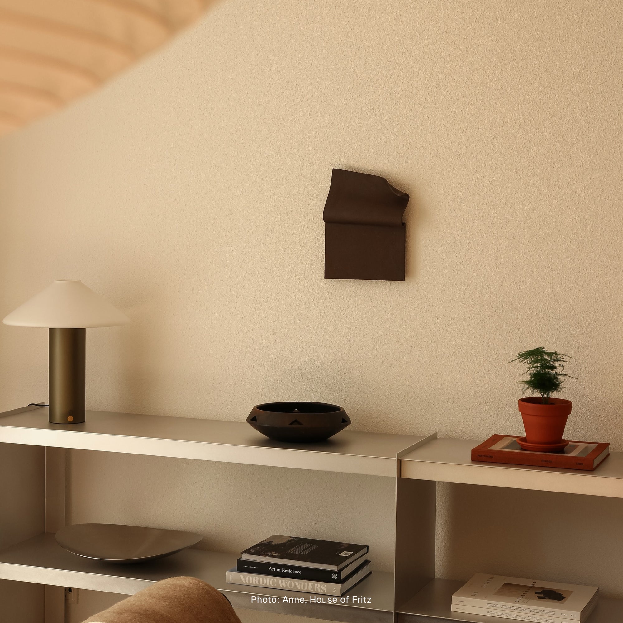 Writings 001 by Studio JOA — Dark Brown folded stoneware wall piece mounted above a shelf styled with a table lamp, ceramic bowl, potted plant and books