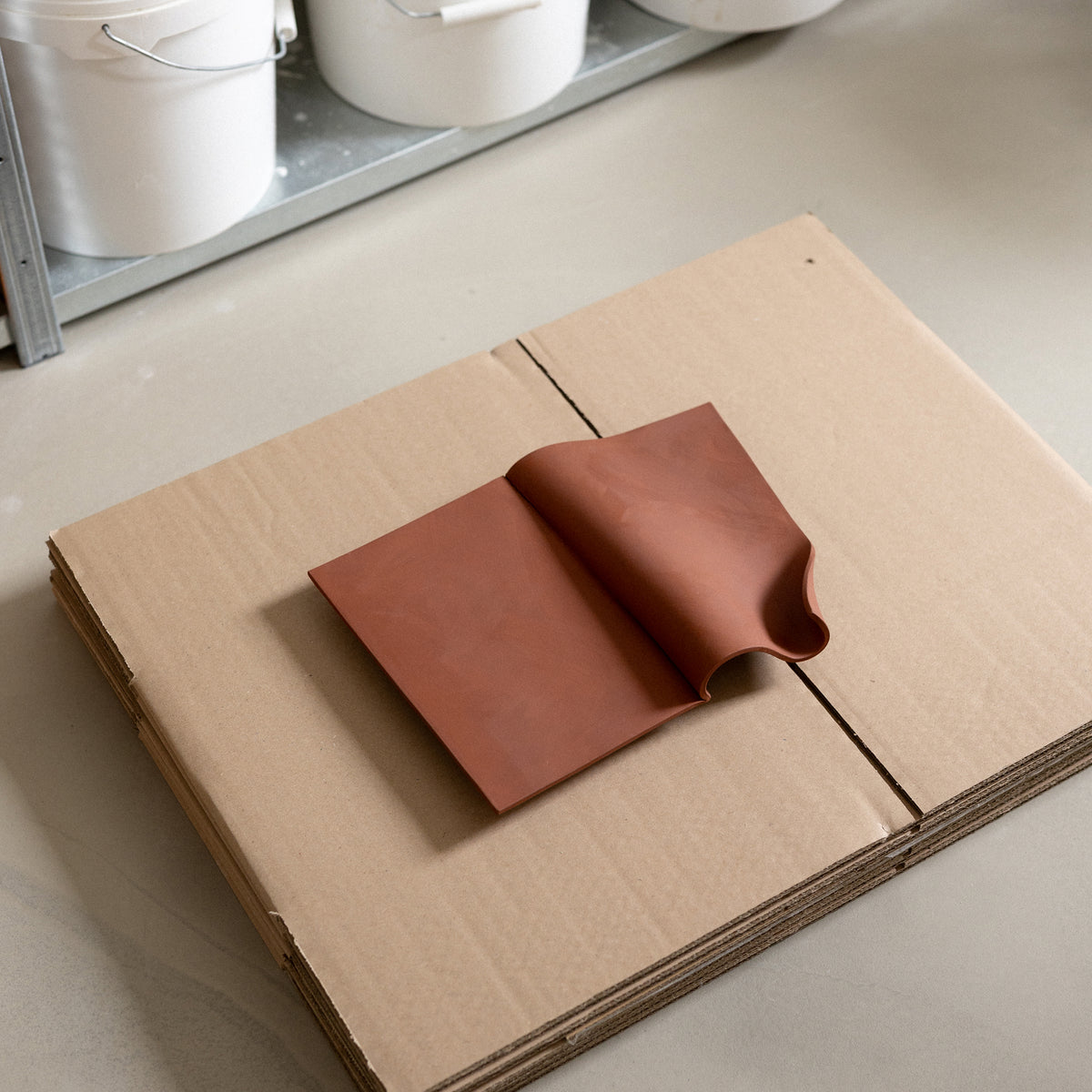 Studio JOA — Stone Red folded stoneware wall piece resting on cardboard in the studio, materials visible in the background