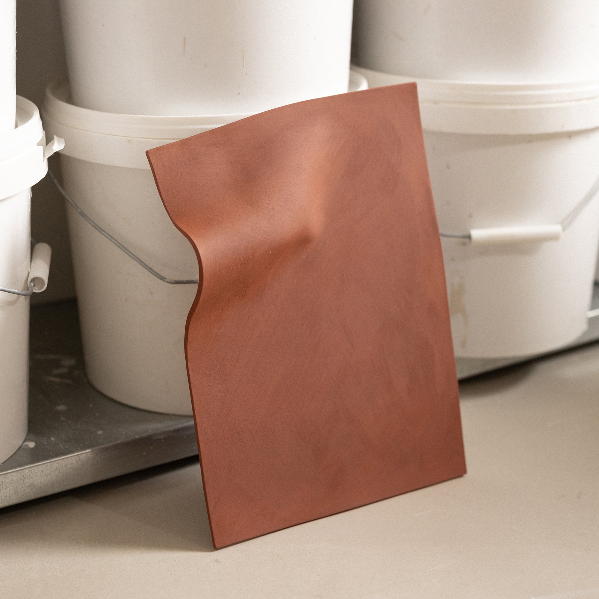 Studio JOA — Stone Red stoneware wall piece standing upright in the studio, leaning against storage buckets